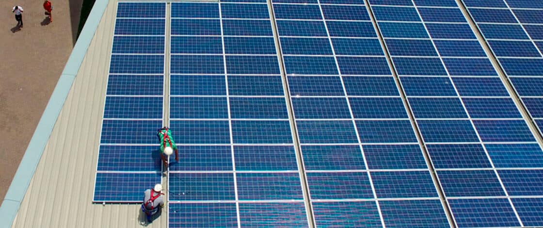 Vista aerea de dos operarios con arnes instalando paneles fotovoltaicos sobre una cubierta metalica.