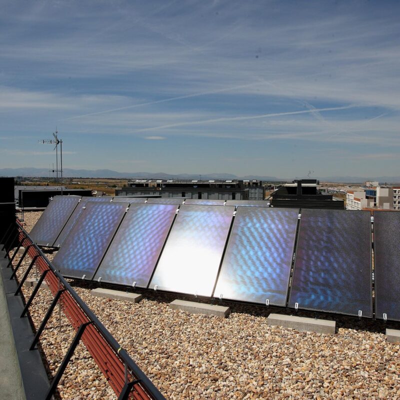 Fila de captadores solares sobre la cubierta plana de un edificio de viviendas, con mas bloques y una grua al fondo